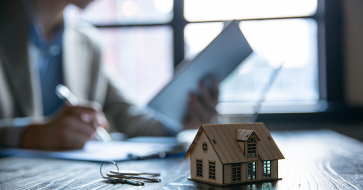 In the background, a person works on mortgage loans at a desk. In the foreground, a small house and a key sit on the desk.In the background, a person works on mortgage loans at a desk. In the foreground, a small house and a key sit on the desk.